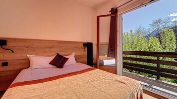 Un dormitorio con cabecera de madera, cama con ropa de cama blanca y una manta estampada, y un balcón con vistas a zonas verdes y montañas.