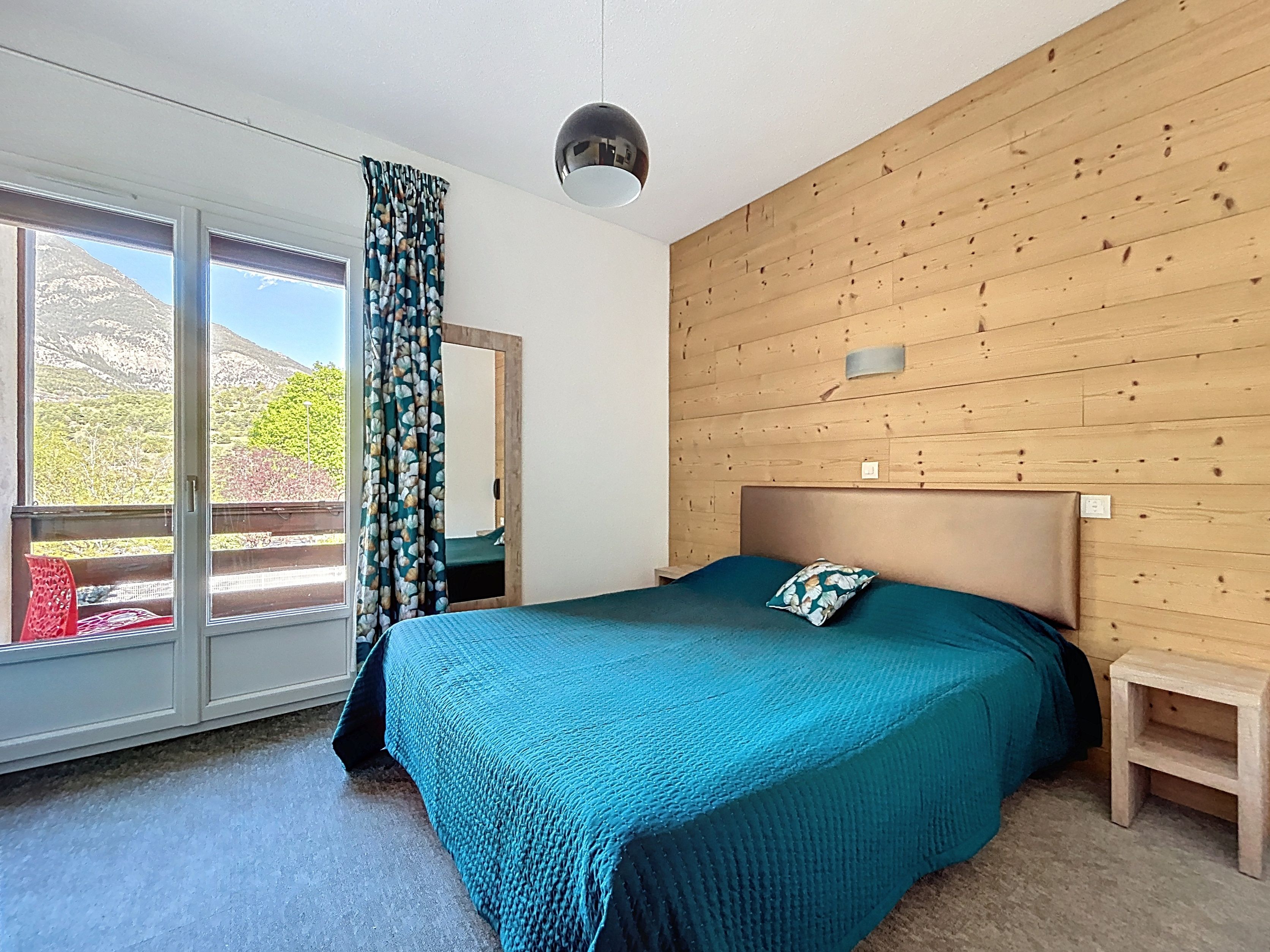 A bedroom with a bed, a small bedside table, a window with curtains, and a view of a mountainous landscape.