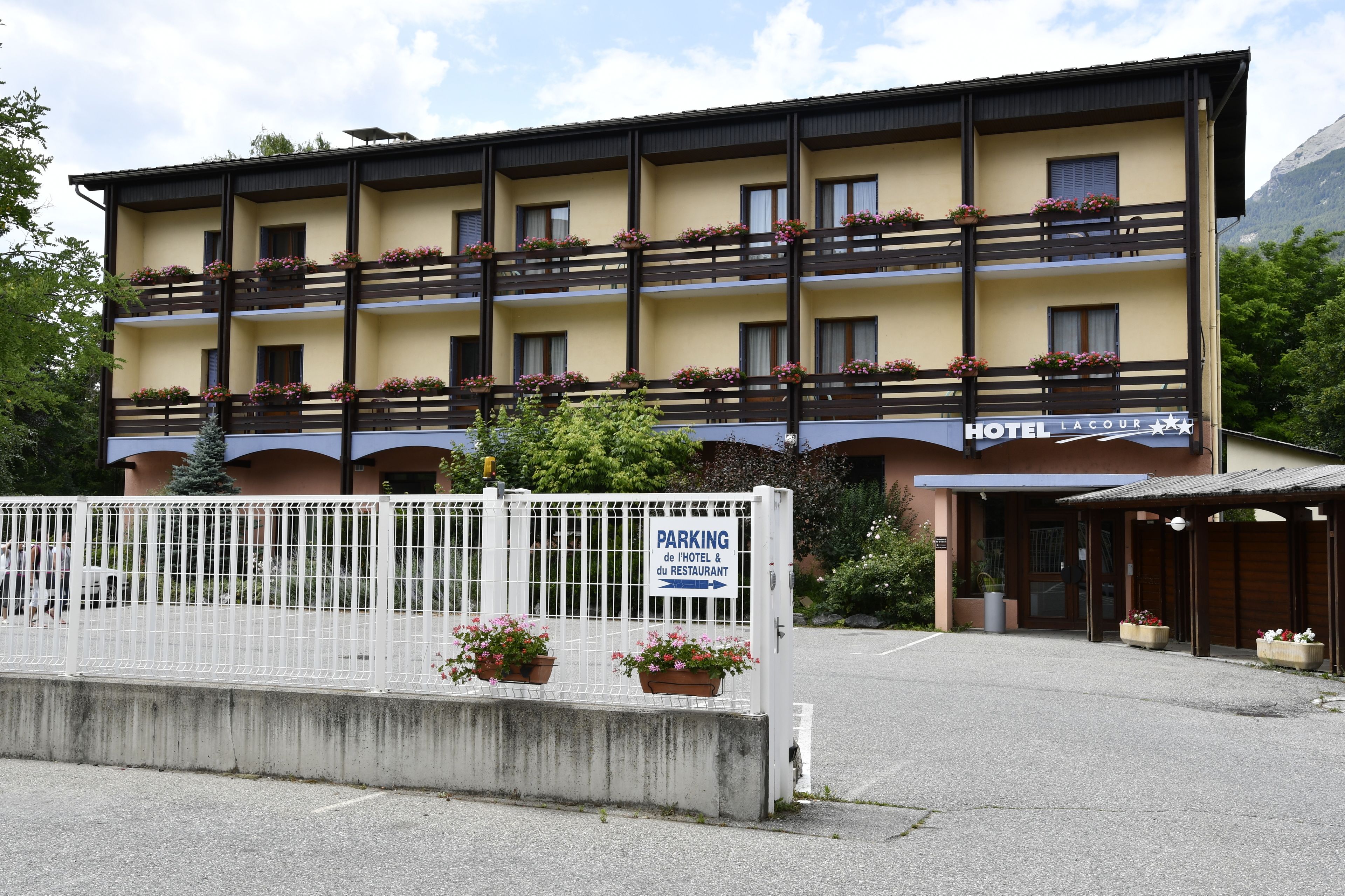 A multi-story hotel with balconies adorned with flowers, a sign indicating parking for the hotel and restaurant, and a mountainous backdrop.