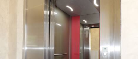 Un ascensor con puertas metálicas y una pared roja visible a través del cristal.