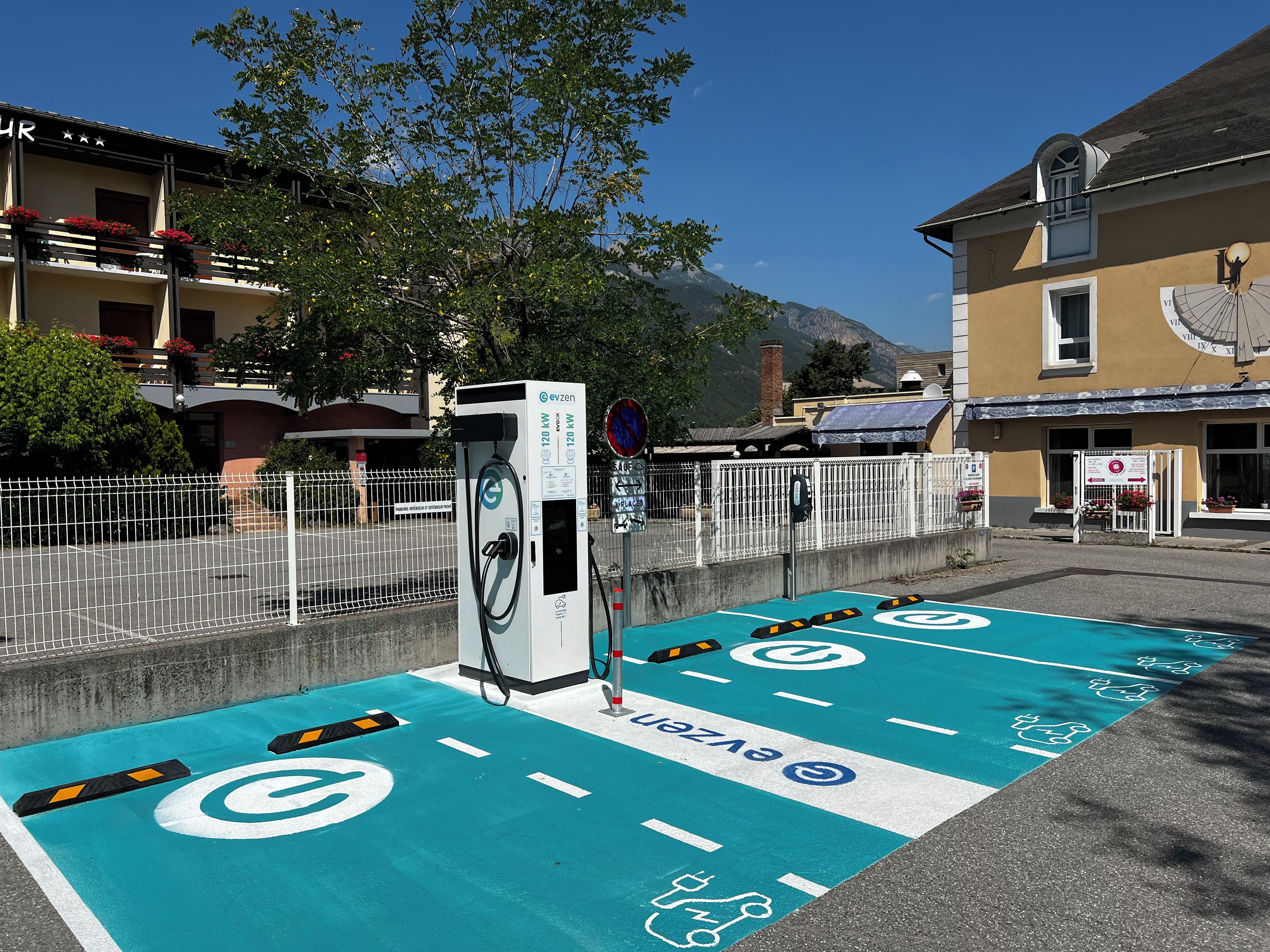 An electric vehicle charging station with a blue parking space, a white fence, and a building with a clock and flower boxes.