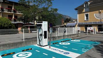 An electric vehicle charging station with a blue parking space, a white fence, and a building with a clock and flower boxes.