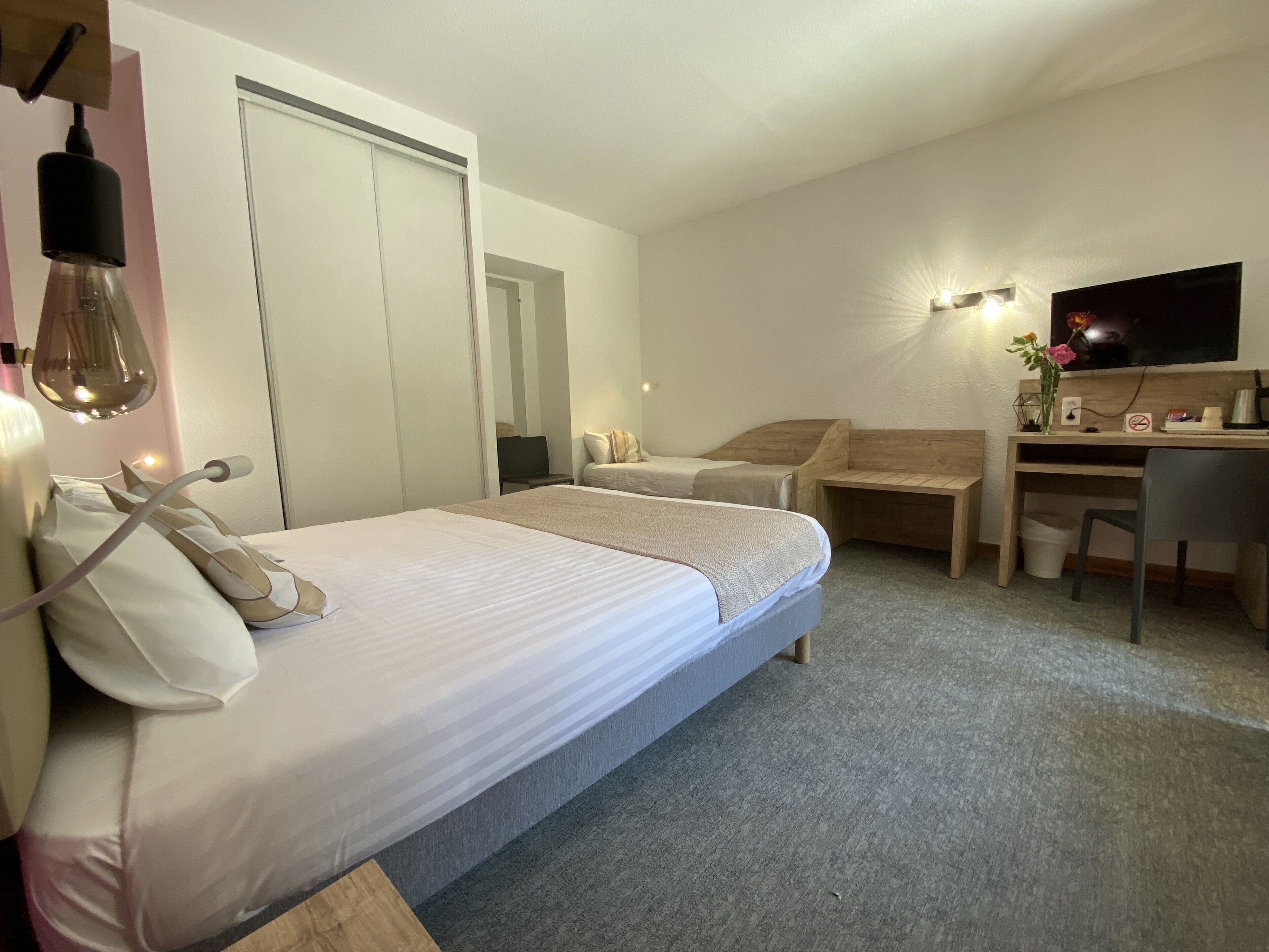 A hotel room with a bed, a desk, a chair, a TV, and a small table with a vase of flowers.