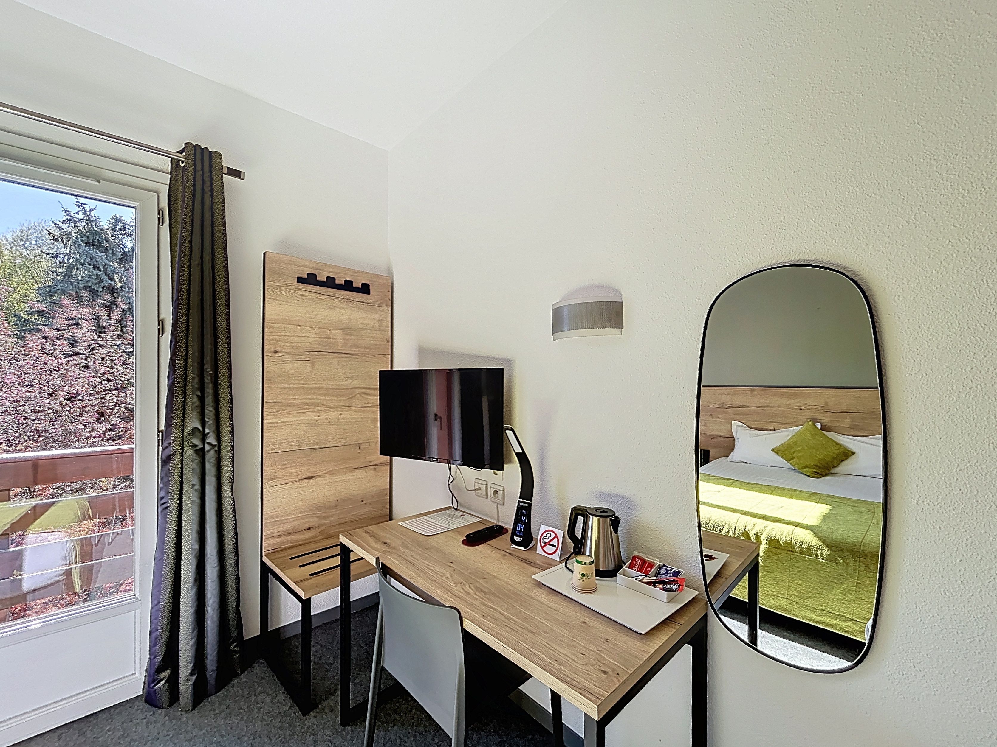 Een hotelkamer met een houten bureau, een flatscreen televisie, een nachtkastje met een waterkoker en een spiegel die de kamer reflecteert.