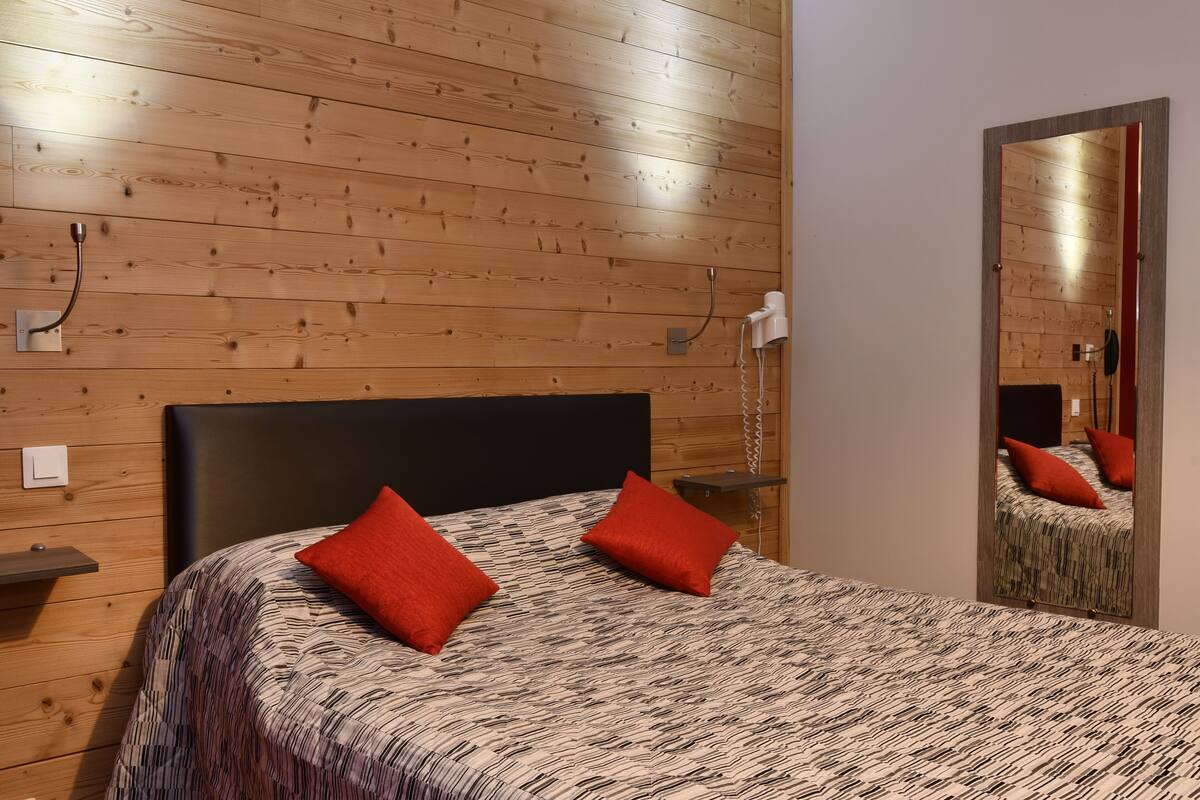 Een slaapkamer met een houten wand, een zwart hoofdeinde, en een bed met een beddengoed met een patroon en twee rode kussens.