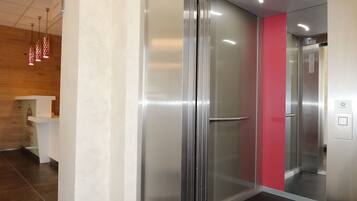 Un ascensor moderno con detalles metálicos y puerta de cristal, que da acceso a un espacio con pared de madera y mostrador blanco.