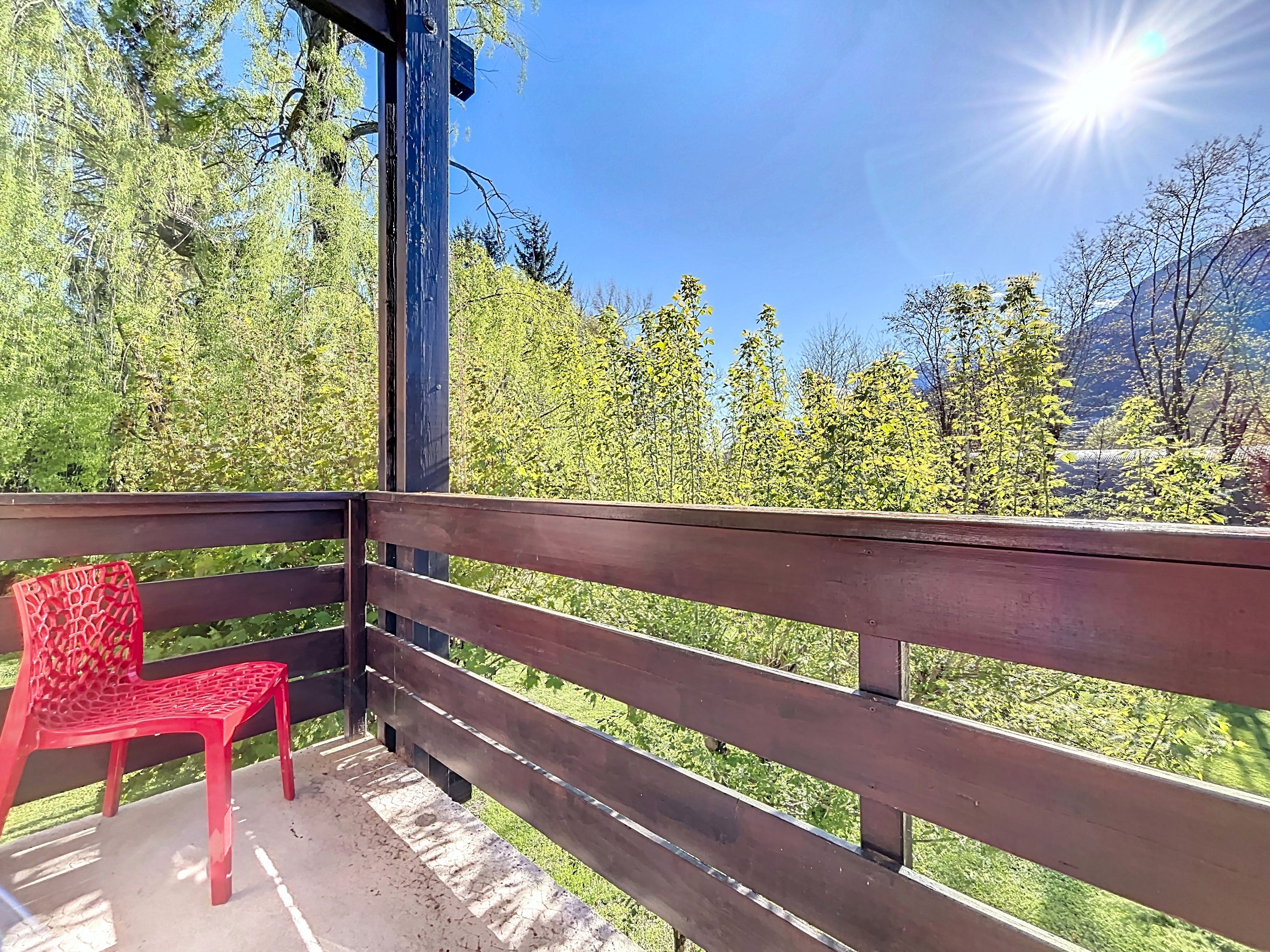 A wooden balcony with a red plastic chair, overlooking a mountainous landscape with green trees.