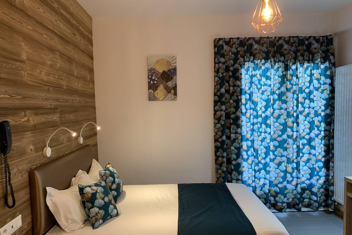 Een slaapkamer met een houten hoofdbord, een bed met blauw-witte bloemenkussens, een zwart plaid en een telefoon op de muur, plus een geometrisch lamparmatuur.