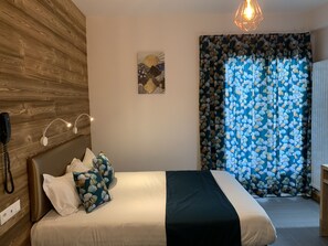 A bedroom with a wooden headboard, a bed with white and blue floral pillows, a black throw blanket, a wall-mounted phone, and a geometric light fixture. - Hotel Lacour (Eygliers)