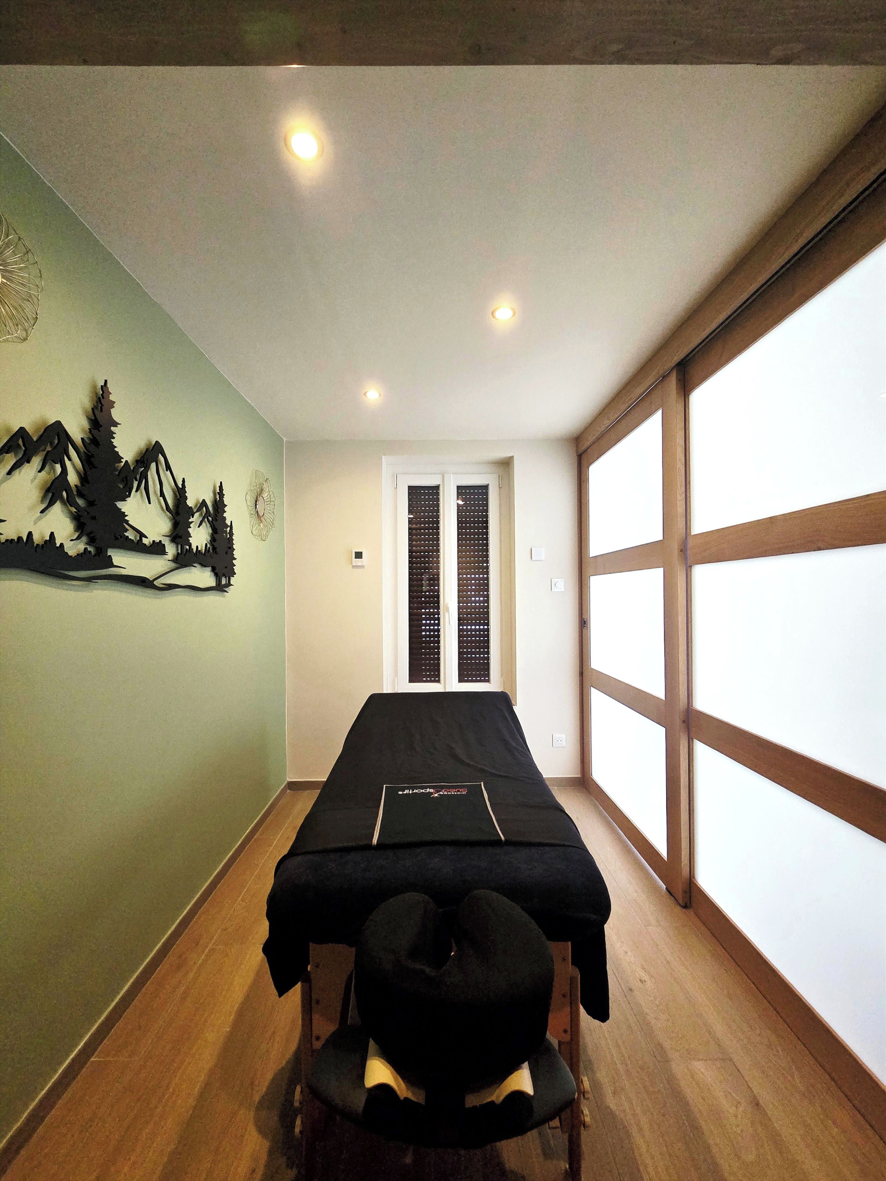 Une pièce équipée d’une table de massage, d’un fauteuil de massage fixé au mur et d’une grande fenêtre avec des stores en bois.