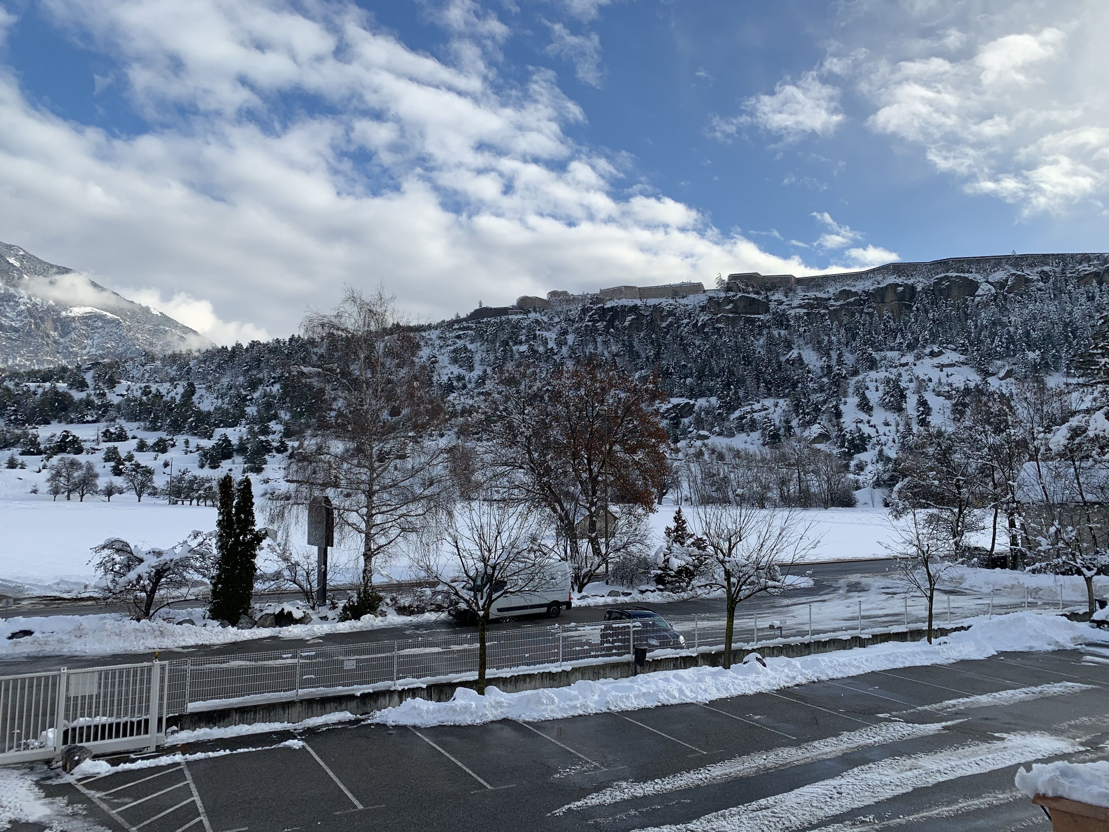 Un estacionamiento con suelo cubierto de nieve, una zona vallada y montañas al fondo.