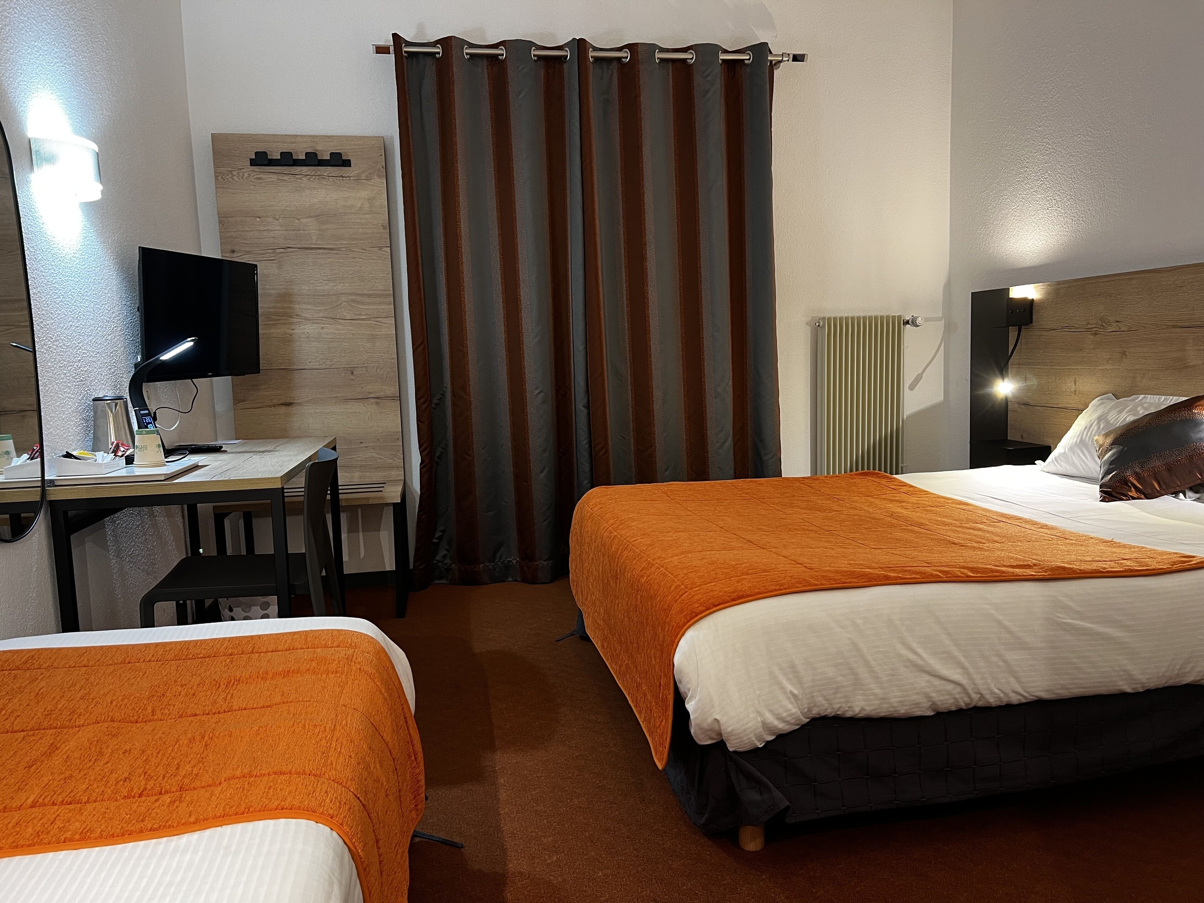 Ein Hotelzimmer mit zwei Betten, einem Schreibtisch mit Computer, einem Stuhl, einem Vorhang und einer Heizung.
