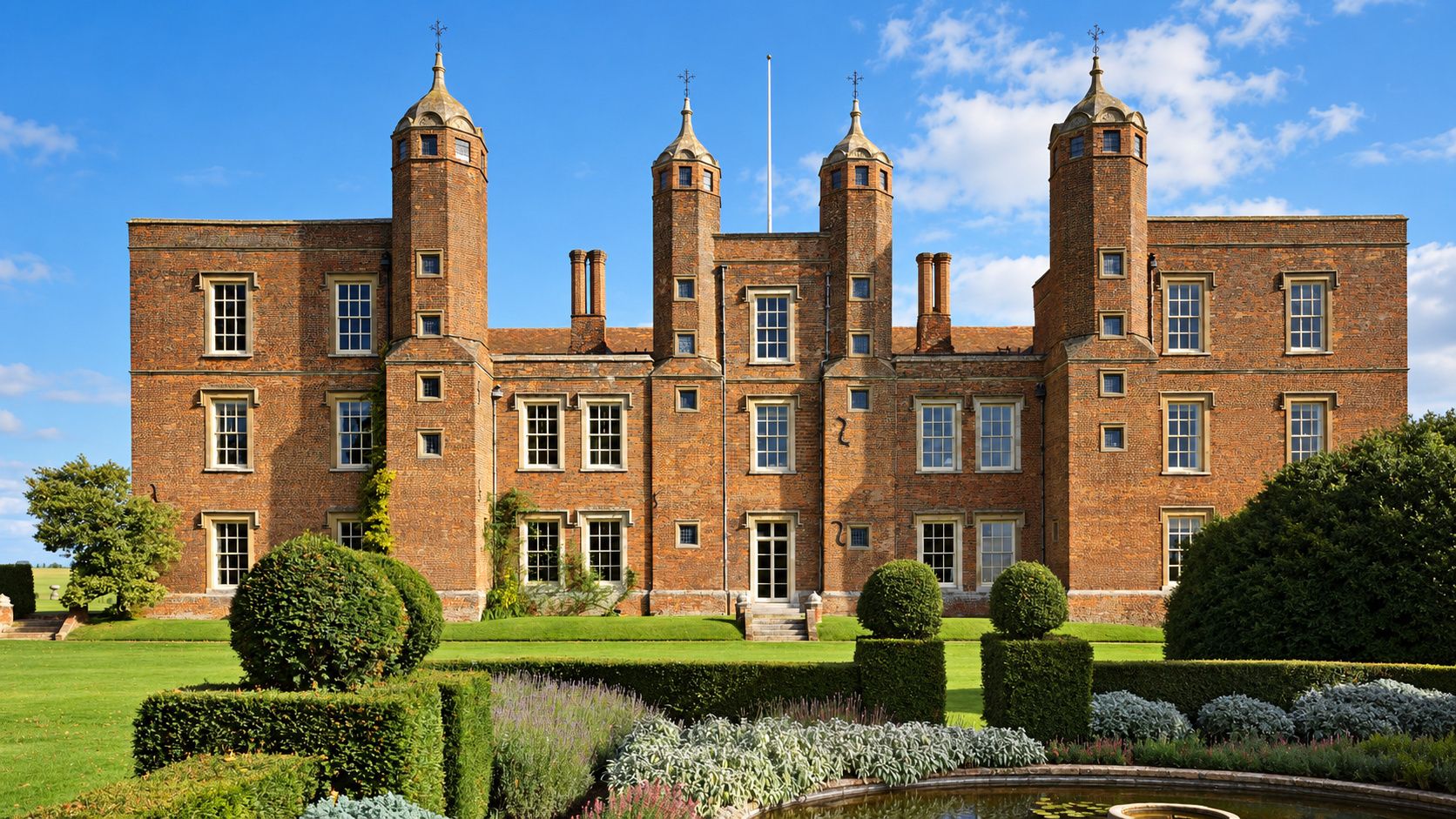 A large brick building with multiple towers and a well-maintained garden with a pond.