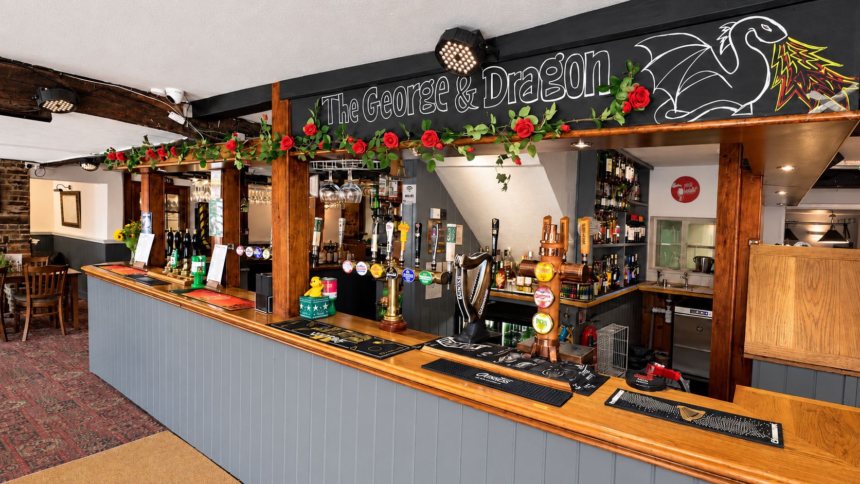 A bar area with a sign reading 'The George & Dragon,' featuring a wooden counter, multiple taps, and a variety of bottles.