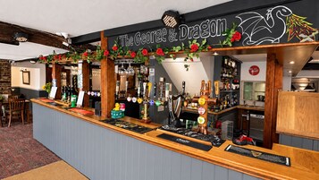 Zona de bar con un letrero que dice 'The George & Dragon', que presenta una barra de madera, múltiples grifos y una variedad de botellas.