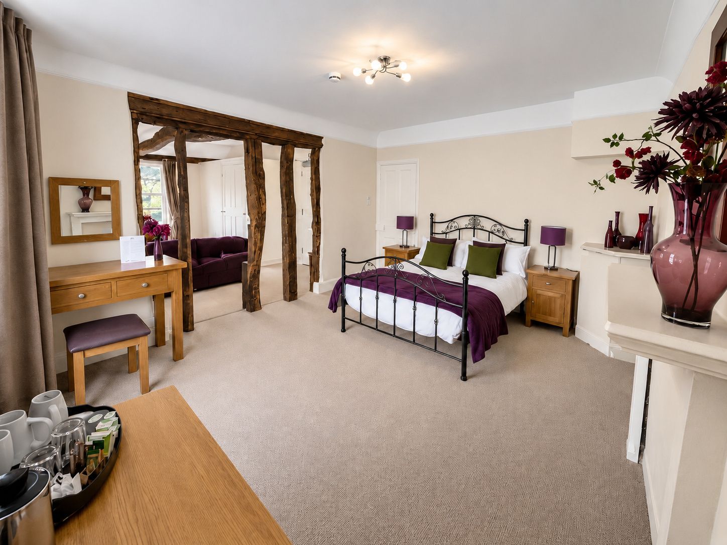 A bedroom with a bed, bedside tables, a desk, a chair, a mirror, and a vase with flowers.