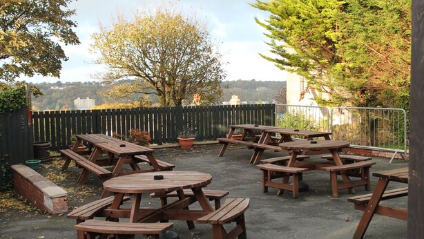 An outdoor patio area with multiple wooden picnic tables and benches, surrounded by a wooden fence and potted plants. - The Menai Hotel and Bar (Bangor)