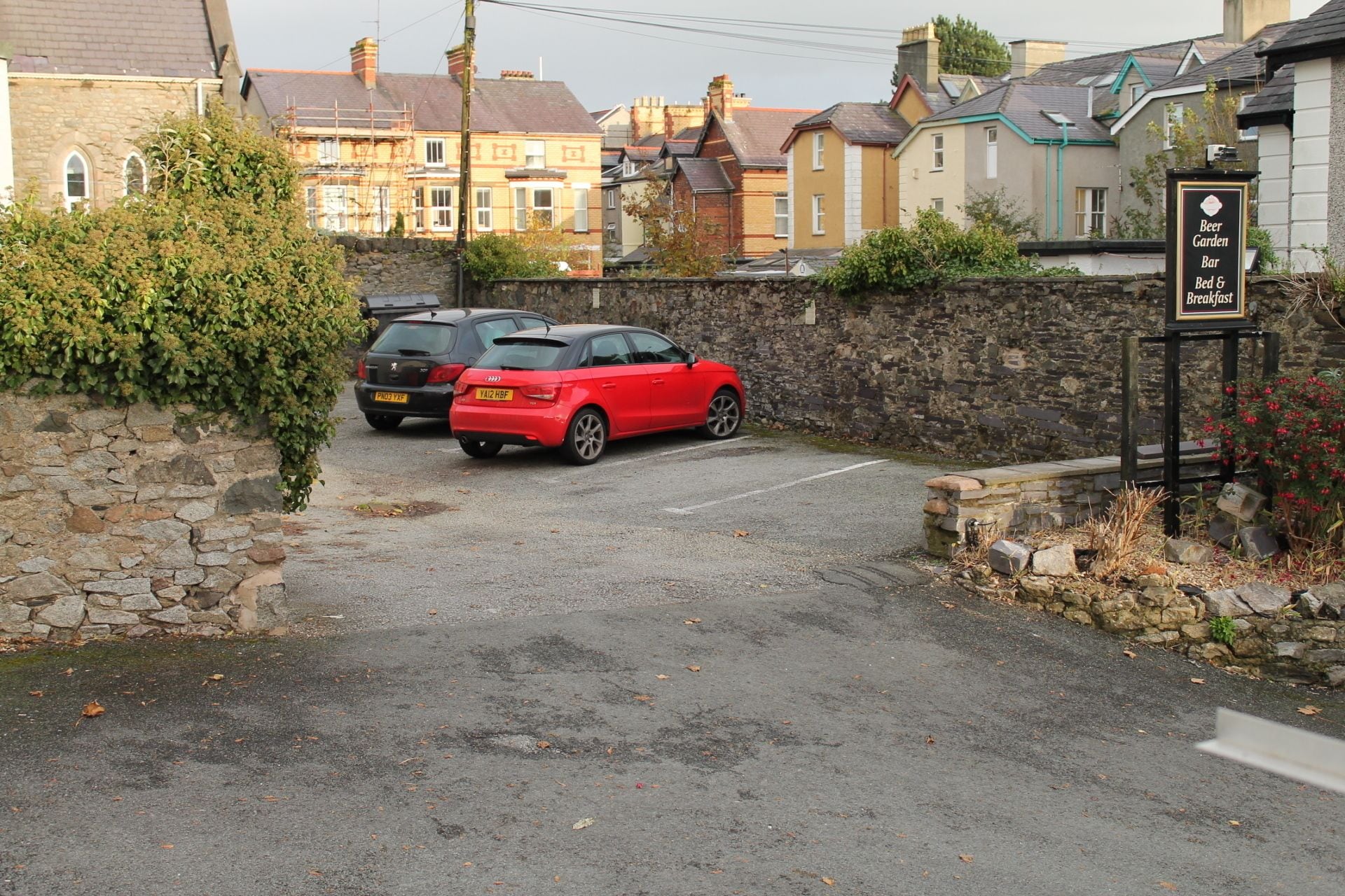 A parking area with two cars, a stone wall, and a building with a sign indicating a beer garden, bar, and bed & breakfast.