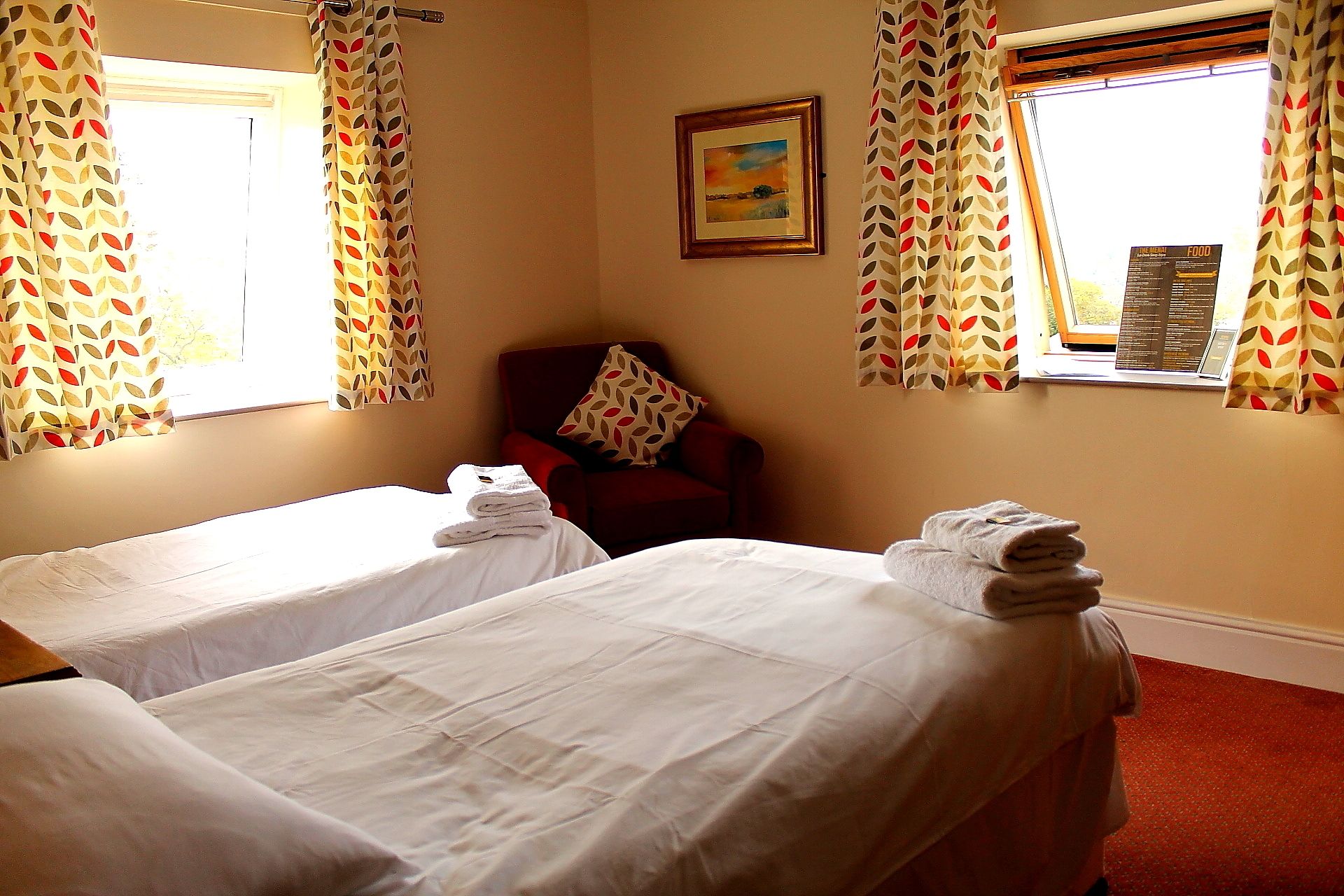 A hotel room with two single beds, a red armchair, and a window with a view of the countryside.