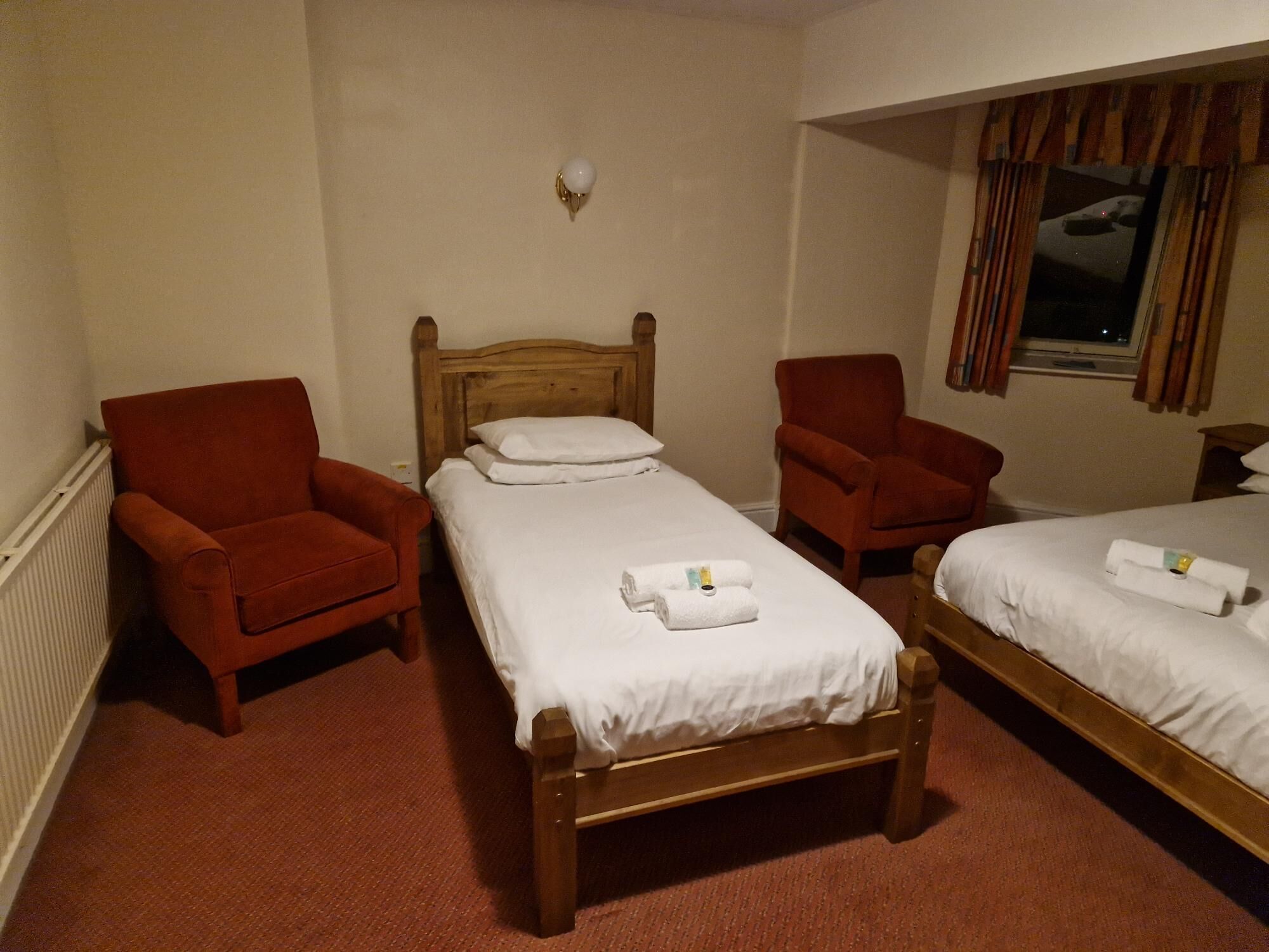 A hotel room with two single beds, a wooden headboard, a red armchair, and a window with curtains.