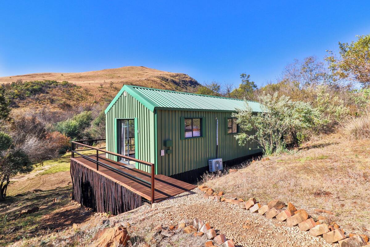 A small, single-story cabin with a green roof and a wooden deck, surrounded by dry grass and shrubs.