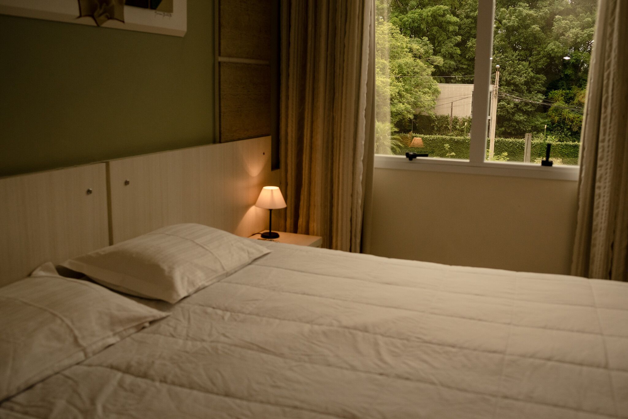 Una cama con ropa blanca, una lámpara de noche y una ventana con vistas a árboles.