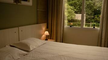 Una cama con ropa blanca, una lámpara de noche y una ventana con vistas a árboles.