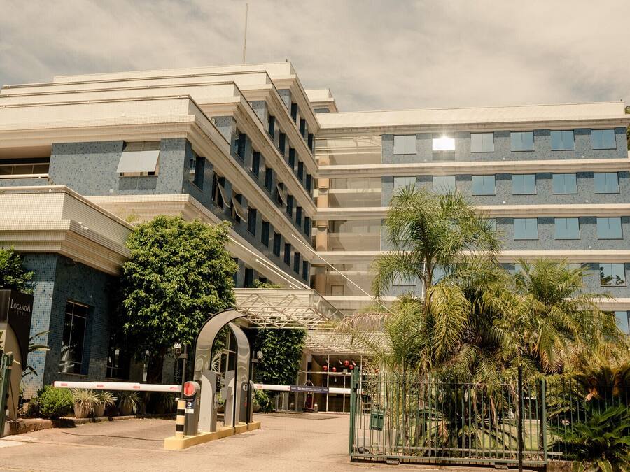 Entrada de un hotel de varios pisos con un camino empedrado, palmeras y cielo despejado.