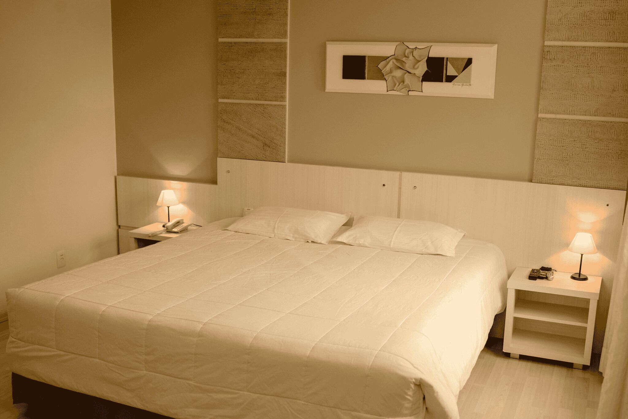 A neatly made bed with white bedding, a bedside lamp, and a small bedside table with a phone and a clock.