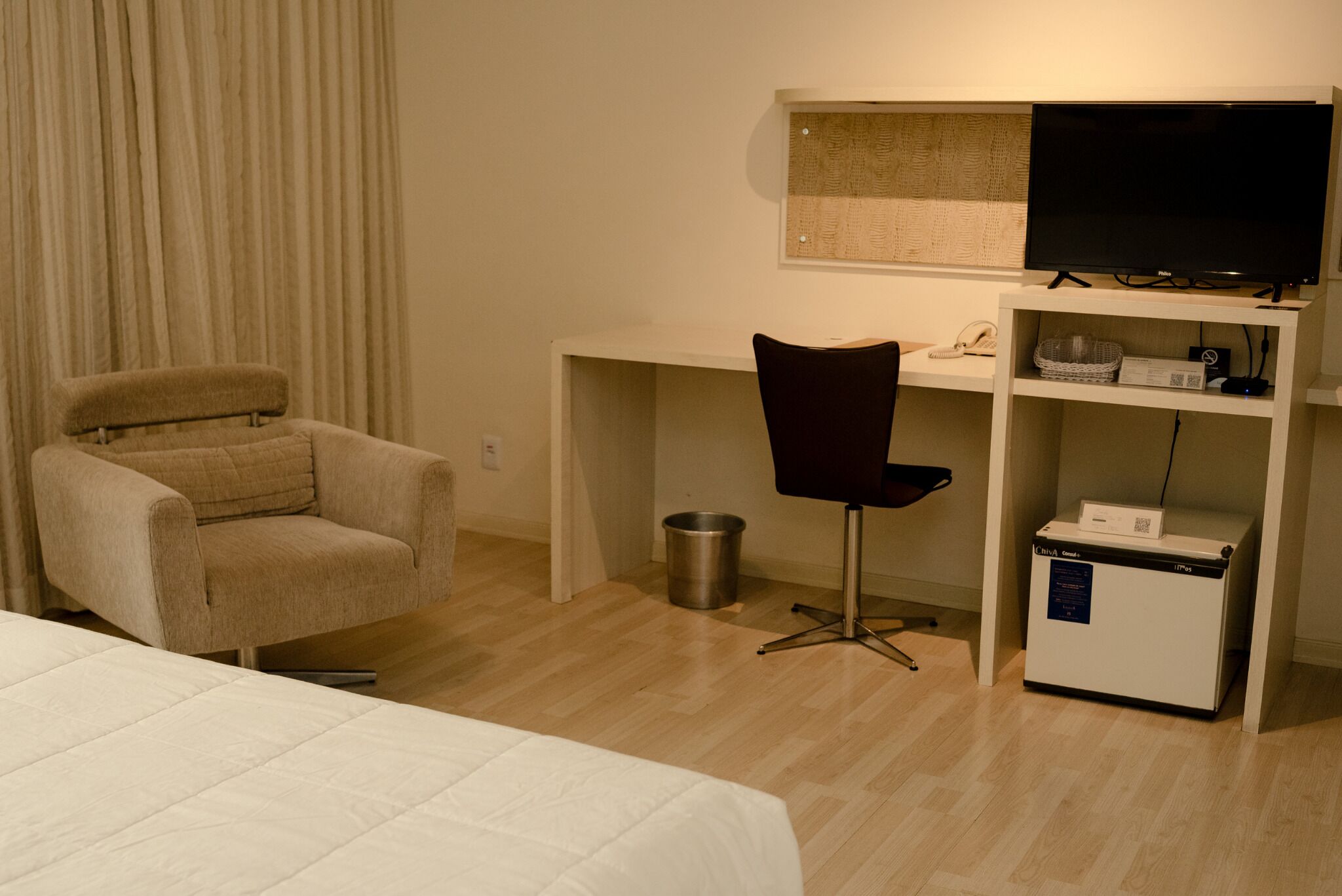 Et hotelværelse med seng, skrivebord, stol, fjernsyn, skraldespand og minibar.