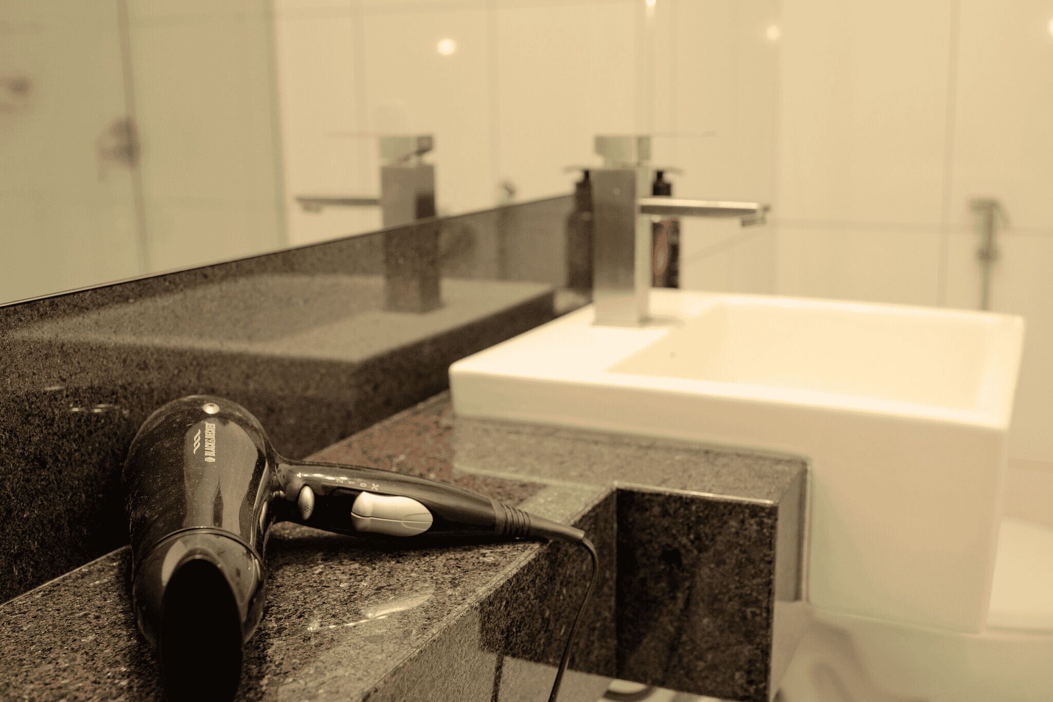 Un sèche-cheveux posé sur un plan de travail en granit dans une salle de bain, avec un lavabo blanc et un miroir qui reflète la pièce.
