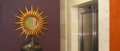 Un ascensor moderno con detalles metálicos, una pared de mármol y un espejo decorativo en forma de sol sobre un console.