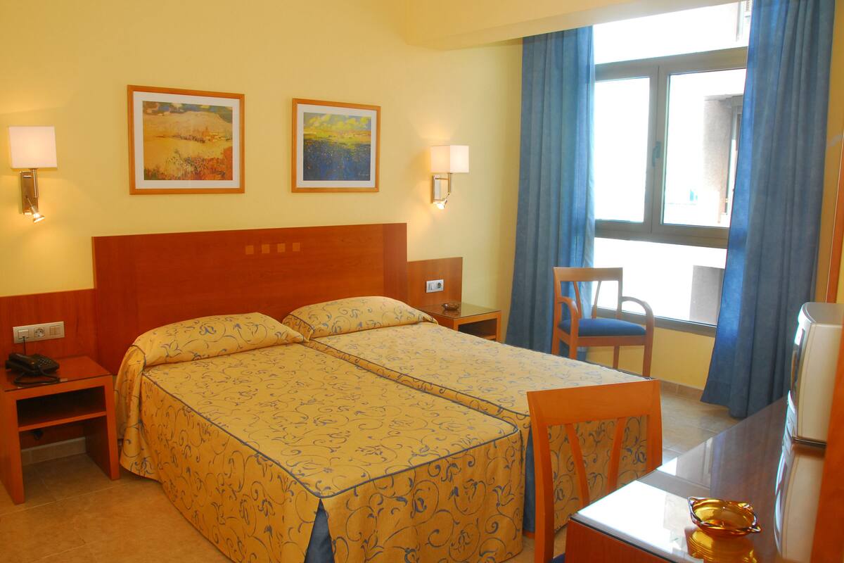 Habitación de hotel con dos camas, un escritorio, una silla, una lámpara y dos cuadros en la pared.