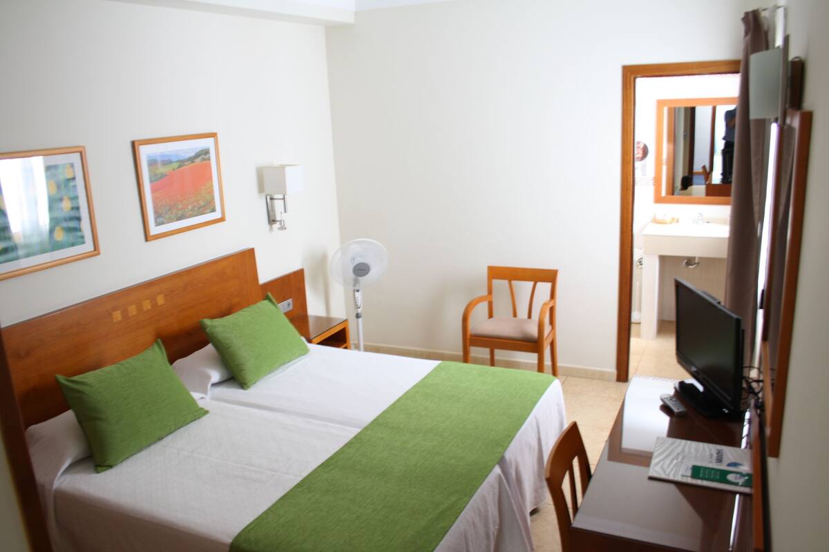 Habitación de hotel con dos camas, una silla, un escritorio con computadora, televisor y obras de arte en las paredes.