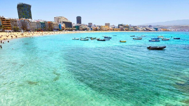 Un paisaje urbano costero con mar azul cristalino, numerosos barcos y una playa animada.