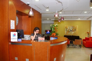 Un área de recepción con un mostrador de madera, un televisor y una zona de estar con sillas naranja y un sofá rojo.