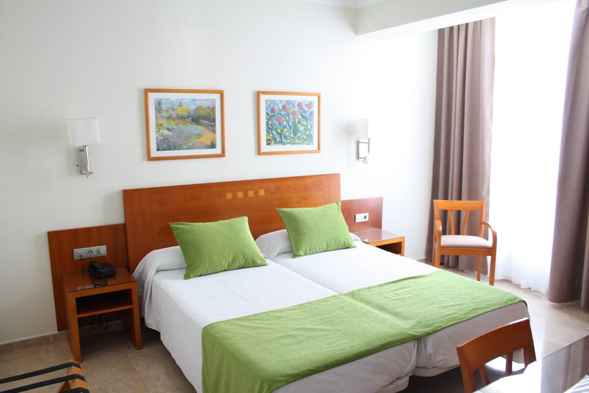 Habitación de hotel con dos camas, almohadas verdes, cabecera de madera, obra de arte enmarcada en la pared, una silla y una mesita de noche con un teléfono.