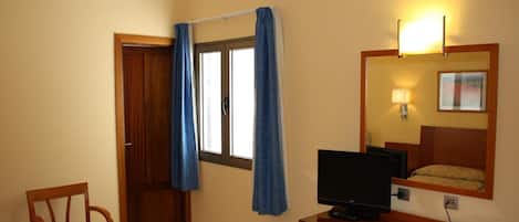 A hotel room with a wooden desk, a television, a chair, a window with curtains, and a mirror with a lamp.