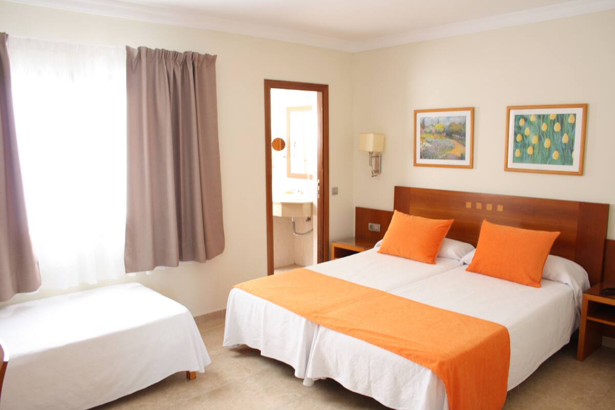Habitación de hotel con una cama grande, una cama más pequeña, una mesita de noche, una lámpara y dos cuadros en la pared.