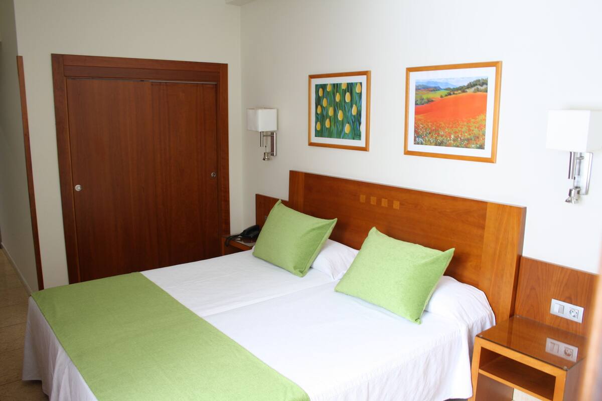 Una cama bien hecha con cubrecama y almohadas verdes, un cabecero de madera, una mesita de noche con una lámpara y dos cuadros enmarcados en la pared.