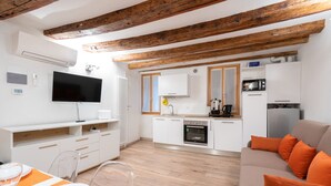A modern kitchen with white cabinets, a built-in oven, and a microwave. There is a dining area with a table set for two, a sofa with orange cushions, and a TV mounted on the wall.