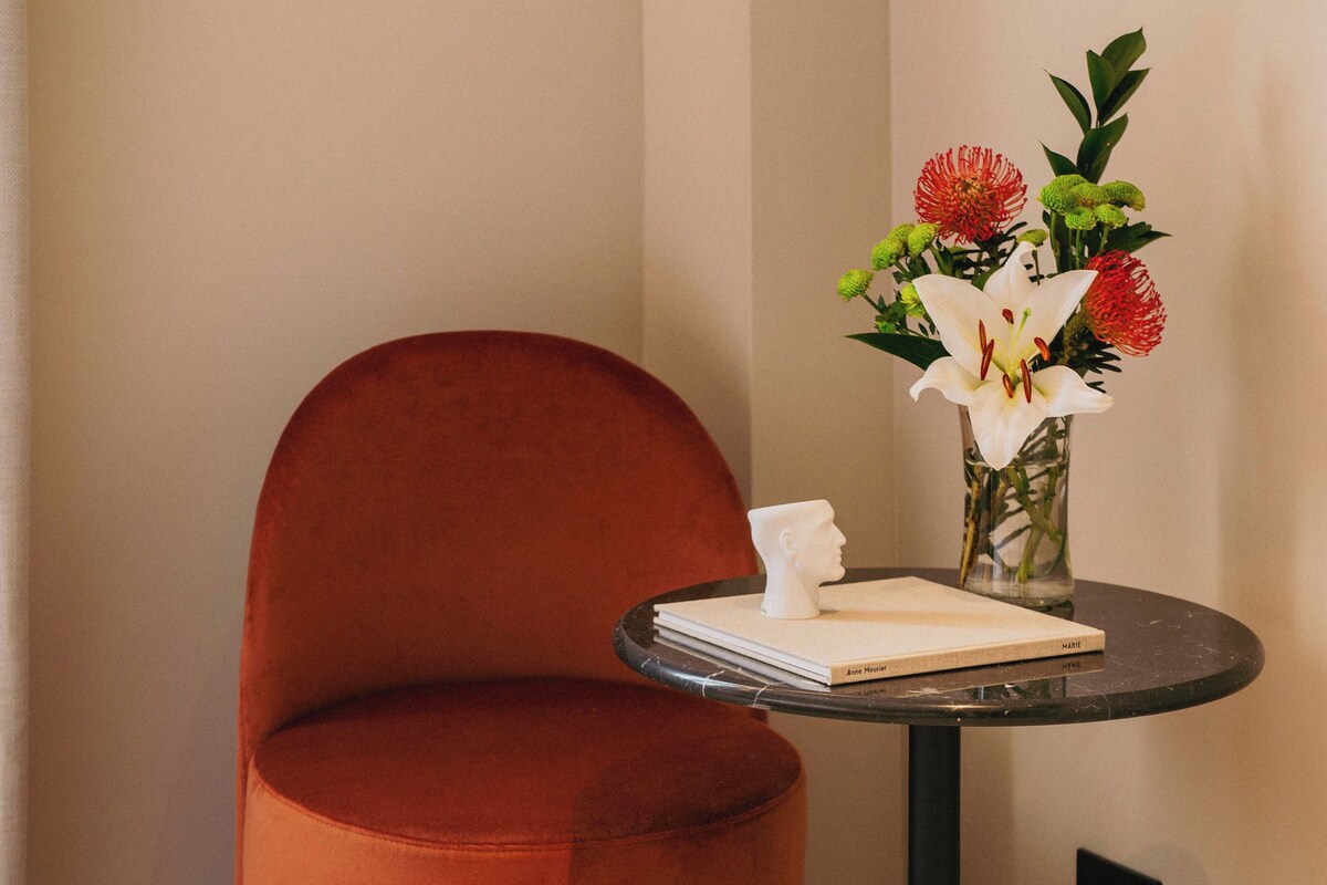 A corner room with a round orange chair, a small round table with a book and a vase of flowers, and a speckled floor.