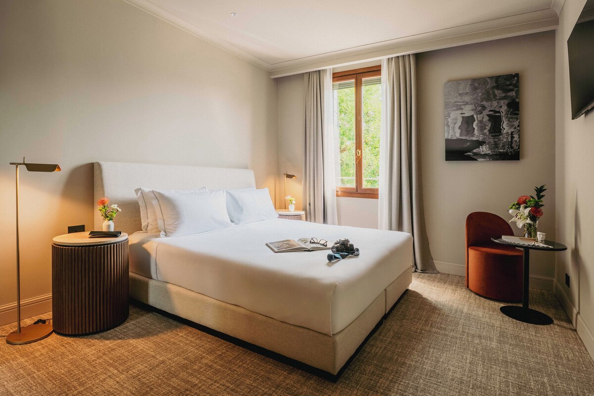 A neatly arranged hotel room with a bed, bedside table, lamp, and a small table with flowers.