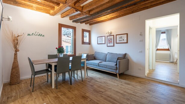 A cozy living room with a wooden ceiling, a grey sofa, a dining table with chairs, and a vase with flowers.
