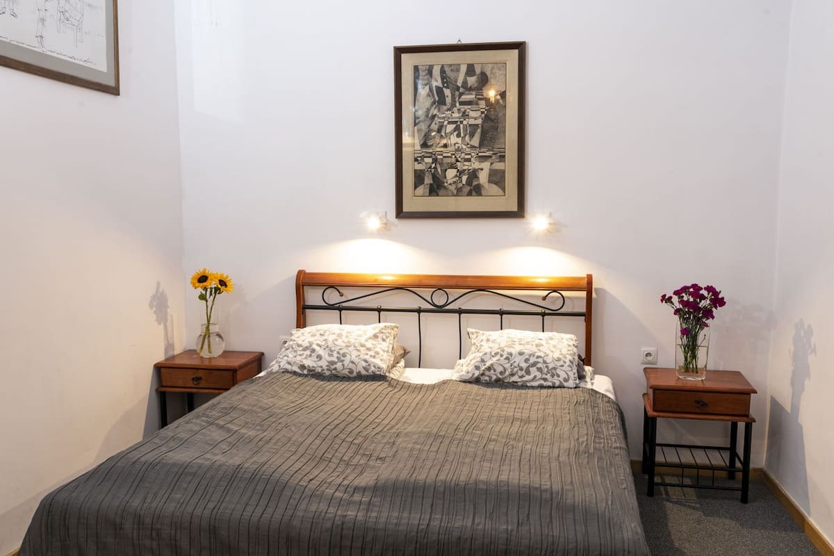 Une chambre bien rangée, avec un lit, deux tables de chevet, une petite table, un tableau encadré au mur et deux vases avec des fleurs.