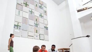 A modern interior with a staircase leading to a second floor, a wall with framed pictures, and a group of people sitting on the steps. - Pathpoint Cologne Backpacker Hostel (Cologne)