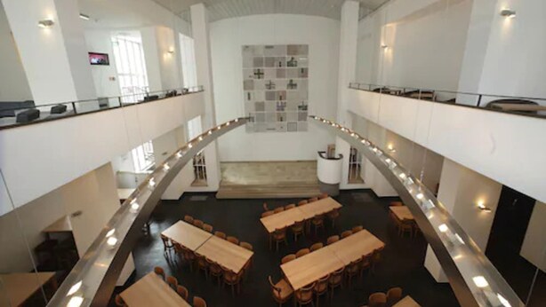 A spacious, modern interior with a central staircase, multiple dining tables, and a wall-mounted display. - Pathpoint Cologne Backpacker Hostel (Cologne)