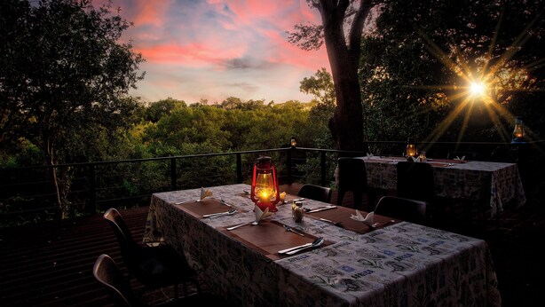 Un área de comedor al aire libre con mesas preparadas para una comida, rodeada de árboles y vistas al atardecer.