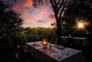 An outdoor dining area with tables set for a meal, surrounded by trees and a sunset view. - Kubu Safari Lodge (Hoedspruit)