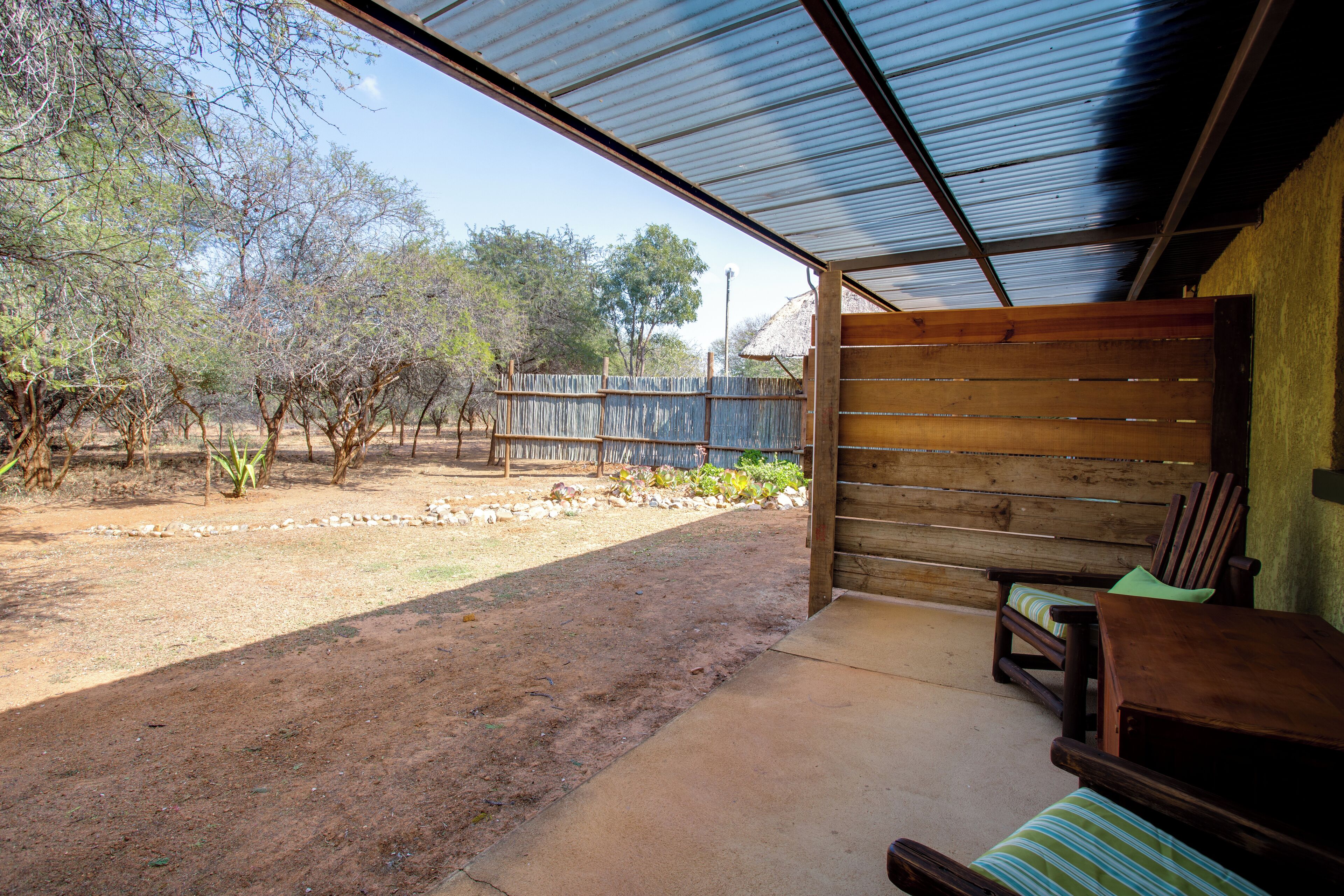 Un espacio al aire libre techado con un banco de madera y una mesa, rodeado por una valla rústica y árboles.